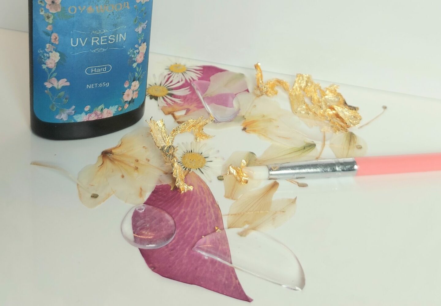 Bouteille de résine UV dure "Ohyauda" de 65g pour la création de bijoux, avec des fleurs séchées, des feuilles d'or et un pinceau, sur un fond fleuri.