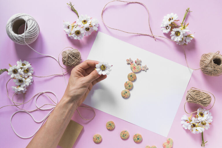 Mains tenant une petite fleur blanche au-dessus d'une feuille de papier vierge entourée de bobines de ficelle, de petites fleurs artificielles et de disques décoratifs faits main sur un fond lilas, illustrant un projet d'artisanat floral.
