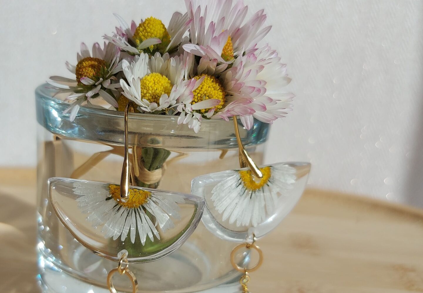 Paire de boucles d'oreilles pendantes artisanales en résine semi-circulaire avec marguerite séchée et perle blanche, posées devant un bouquet de paquerettes dans un vase en verre.
