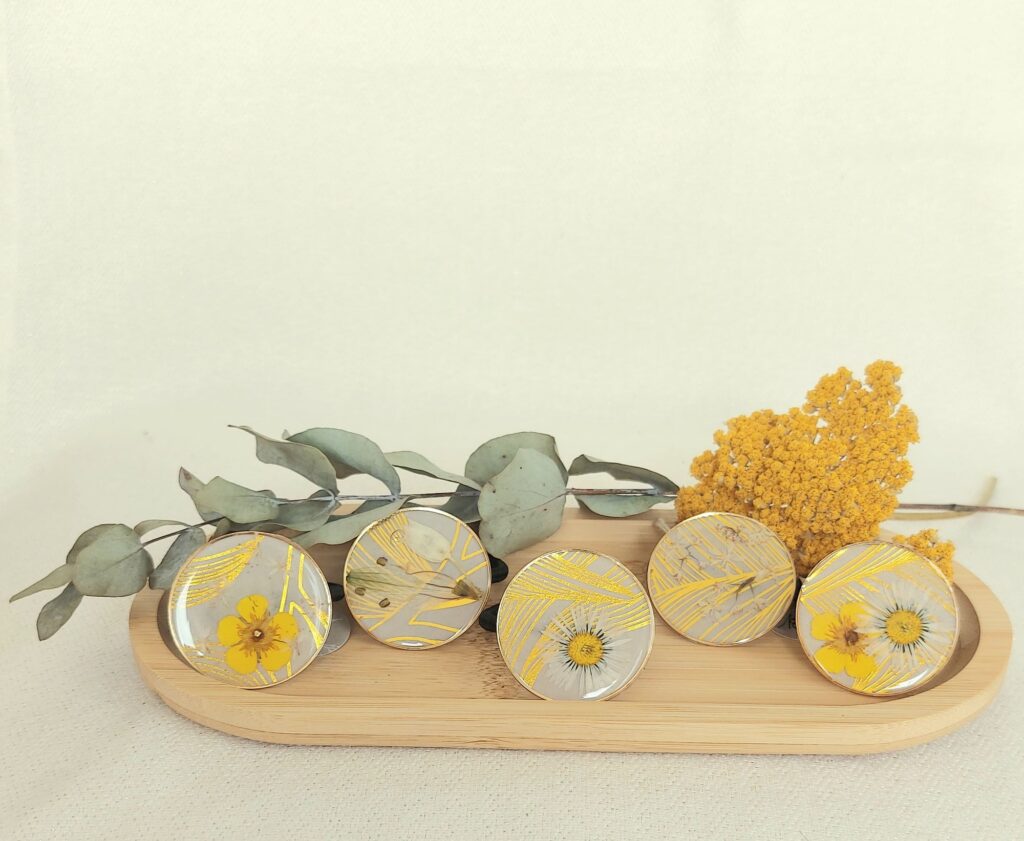 Cinq accessoires de cheveux ronds (élastiques) avec inclusions de fleurs jaunes séchées et feuilles dorées dans de la résine, posés sur un plateau en bambou, avec une branche d'eucalyptus et une fleur d'achillée jaune en arrière-plan.