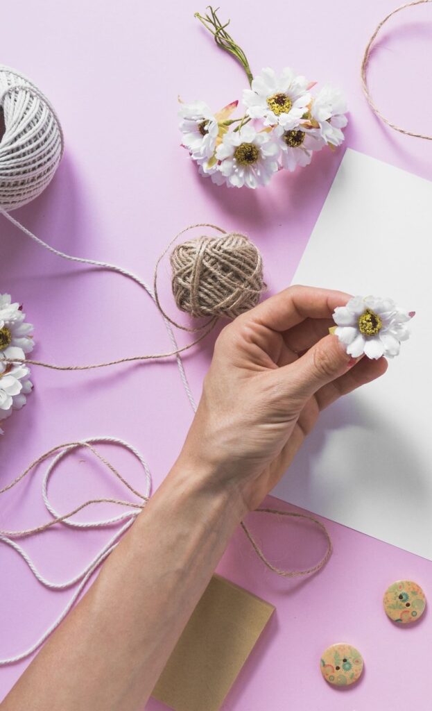 Mains tenant une petite fleur blanche au-dessus d'une feuille de papier vierge entourée de bobines de ficelle, de petites fleurs artificielles et de disques décoratifs faits main sur un fond lilas, illustrant un projet d'artisanat floral.