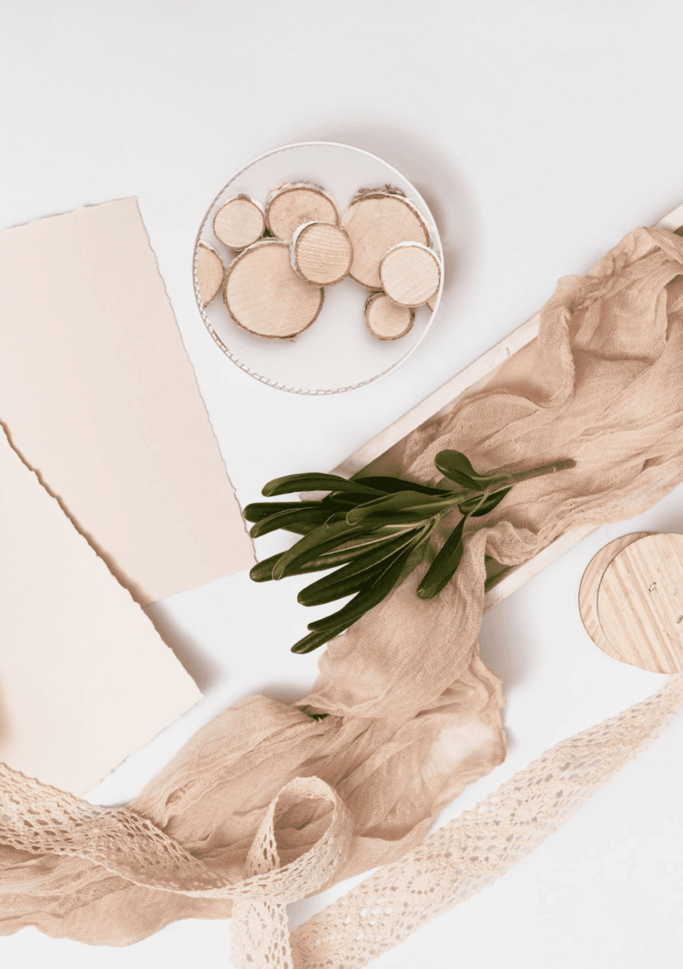 Composition de matériaux naturels et upcyclés incluant des papiers déchirés crème, des rondelles de bois clair, un ruban de gaze froissée beige et de la dentelle vintage sur fond blanc.