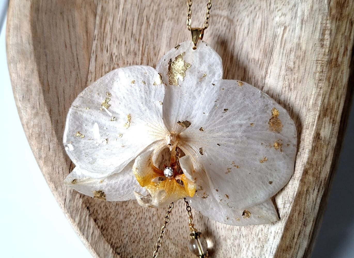 Sautoir artisanal AnneFicelle Créations avec une véritable fleur d'orchidée blanche résinée au pinceau et incrustée de feuilles d'or, posé sur un support en bois.